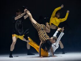 4 dancers pose in a group. One sits with hands and legs in the air. One does a handstand. Two stand.