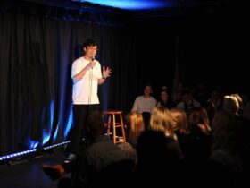 Phil Wang on stage, gesturing outwards, in front of a sold out crowd