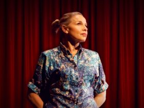 Performer Justine Clarke stands against red theatre curtain, looks to the side