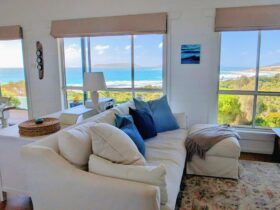 White sofa with blue cushions, lamp and large windows look out to ocean view.
