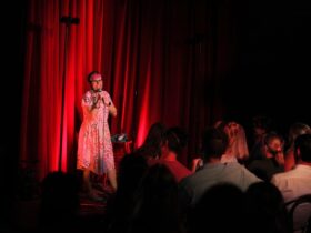 Cal Wilson performing on stage against a bright red curtain. There is a seated crowd in front